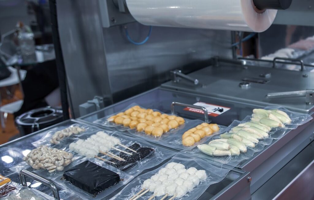 Thermoforming packaging machine sealing assorted food trays with clear plastic film.